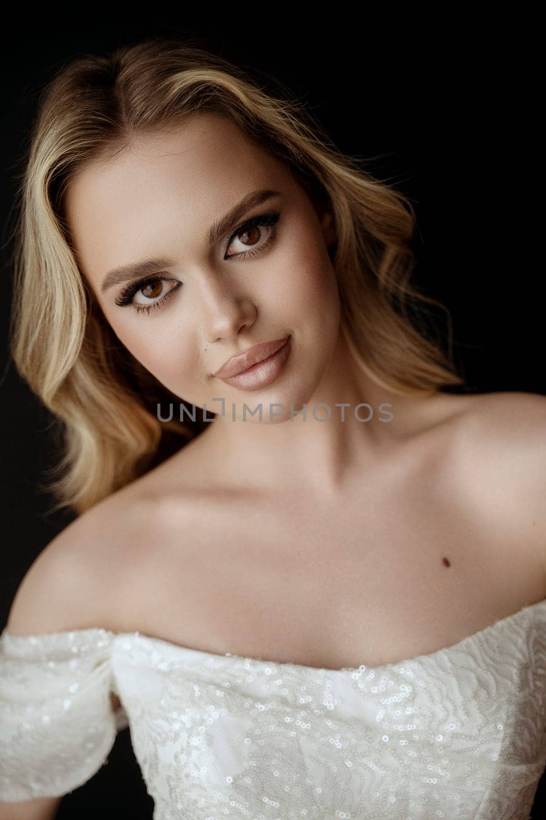 Portrait Young, fair-haired woman with wavy hair. Beautiful model with long hairstyle. Blonde with perfect makeup. In a white dress with open shoulders. On a dark background looks at the camera