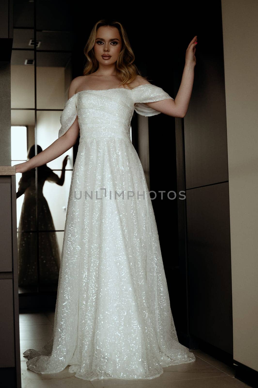 Elegant wedding dress presented in a minimalist interior with a model in a chic pose. Blonde bride against a dark interior. Bride waiting for the groom. Looking at the camera, shot from a low angle