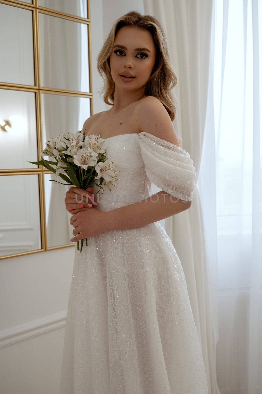 Portrait of a blonde bride with a bouquet of white flowers by the window. Blonde in a flowing wedding dress, radiates grace and beauty. In a light minimalist interior with a window and a mirror