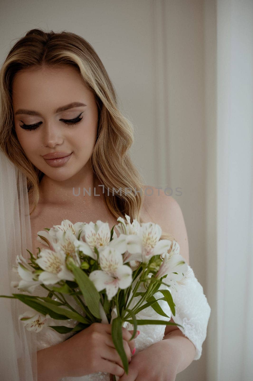 Portrait of a blonde bride with a lush veil, in a white dress with a bouquet of lilies. Close-up on a white background. Bride with makeup and wavy long hair