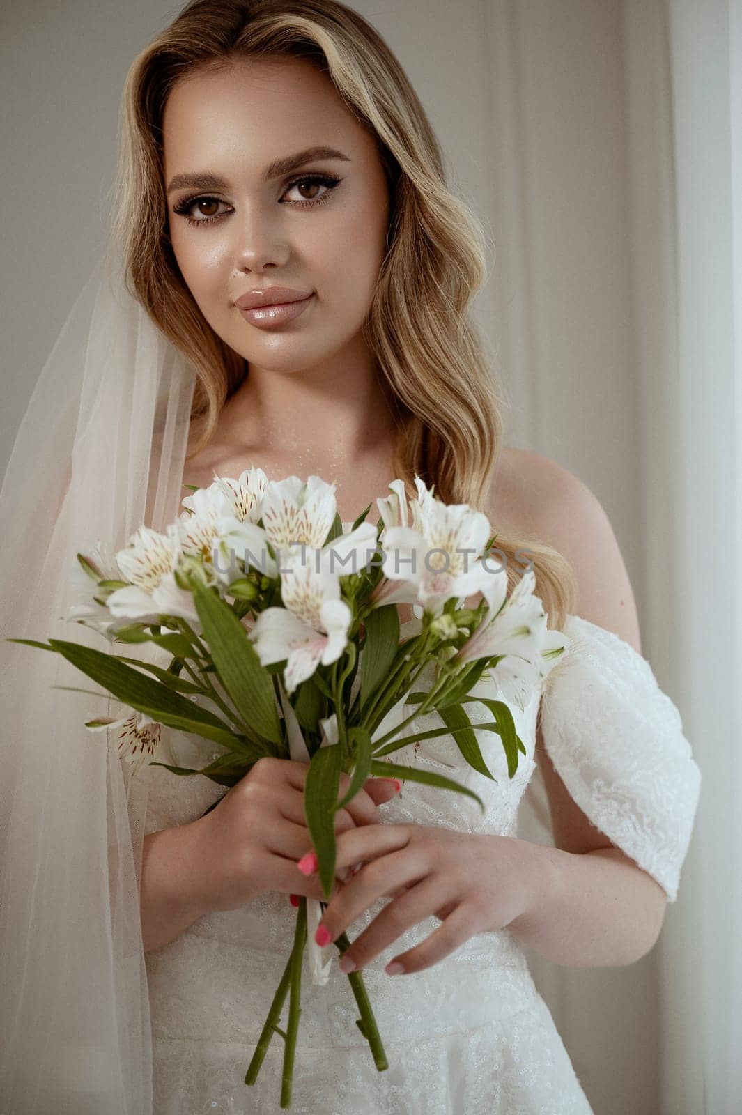 Portrait of a blonde bride with a lush veil, in a white dress with a bouquet of lilies. Close-up on a white background. Bride with makeup and wavy long hair, smiling