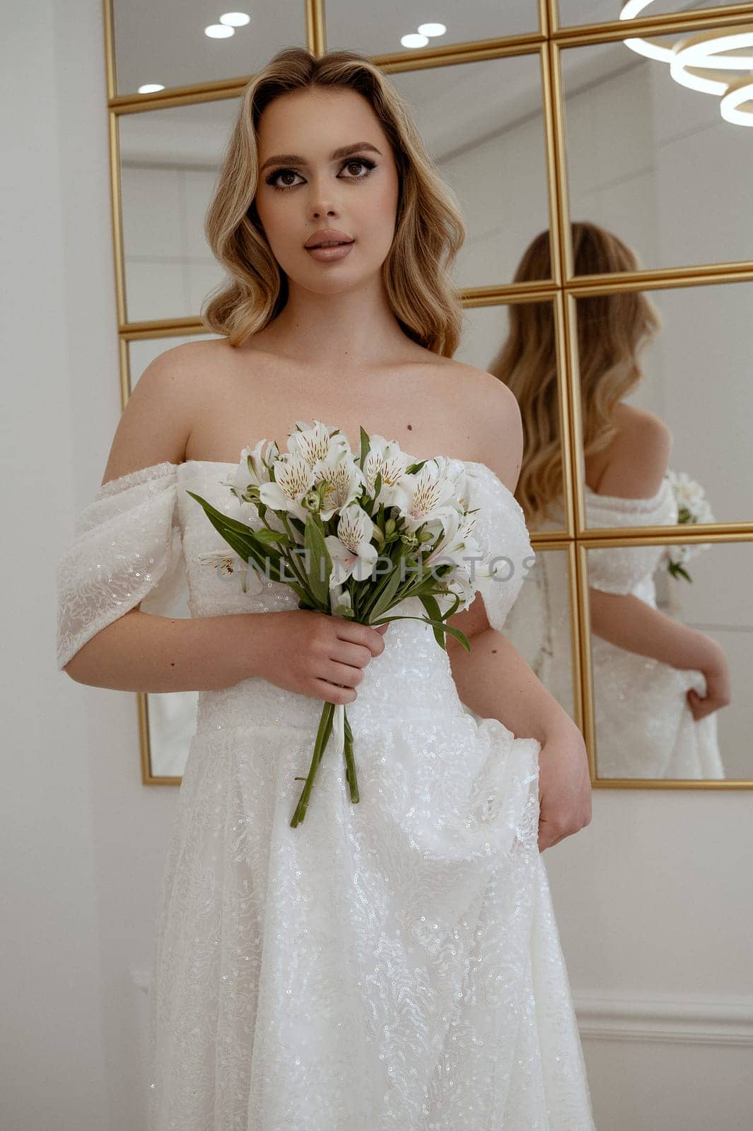 Portrait of a blonde bride with a bouquet of white flowers against the background of a mirror. Blonde in a flowing wedding dress, holding white lilies. In a light interior, looking at the camera