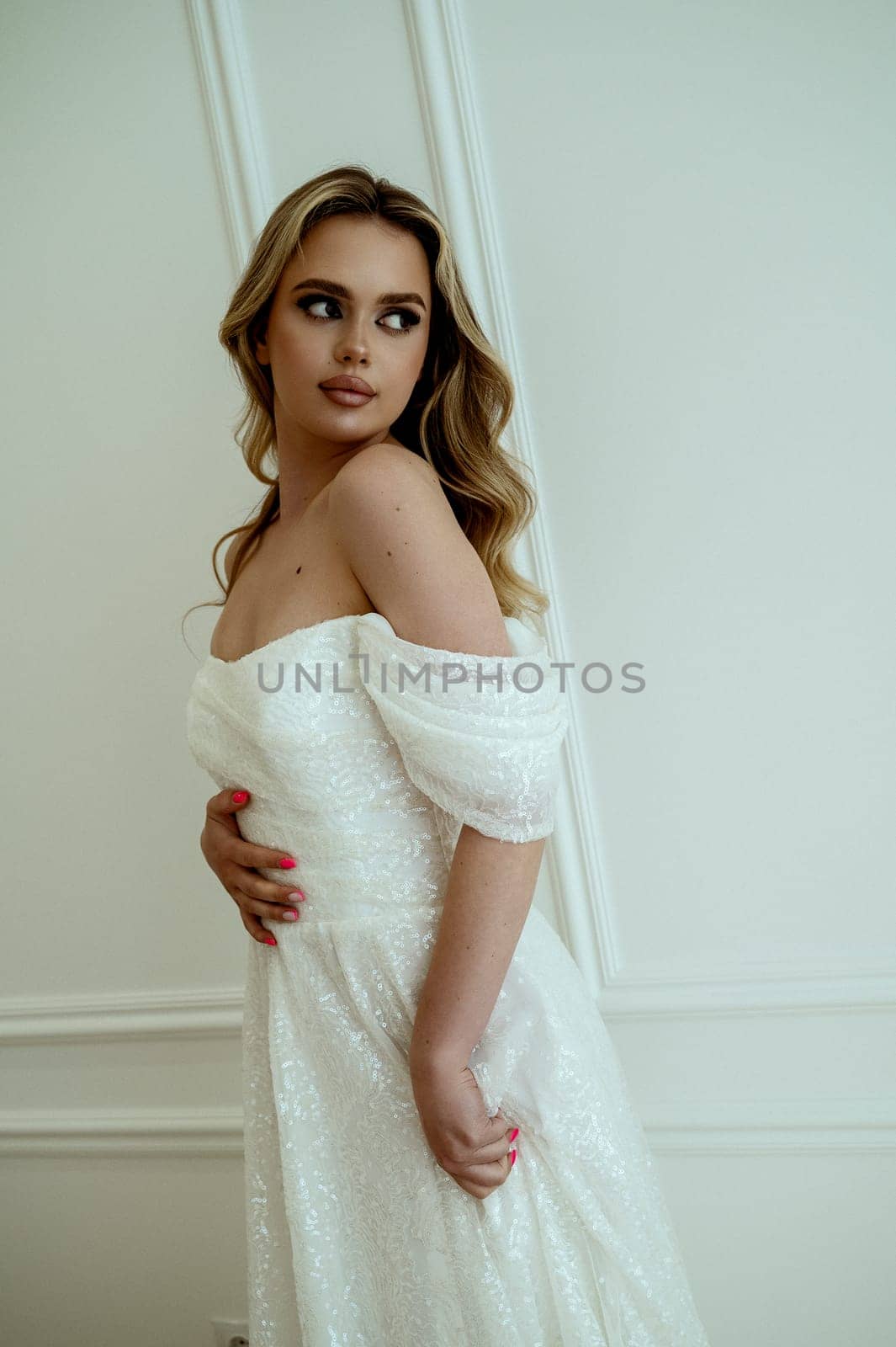 harming bride dynamically poses in flowing wedding dress on white background in minimalist interior. She radiates grace and beauty. Blonde with long hair elegantly poses, profile, styling, makeup