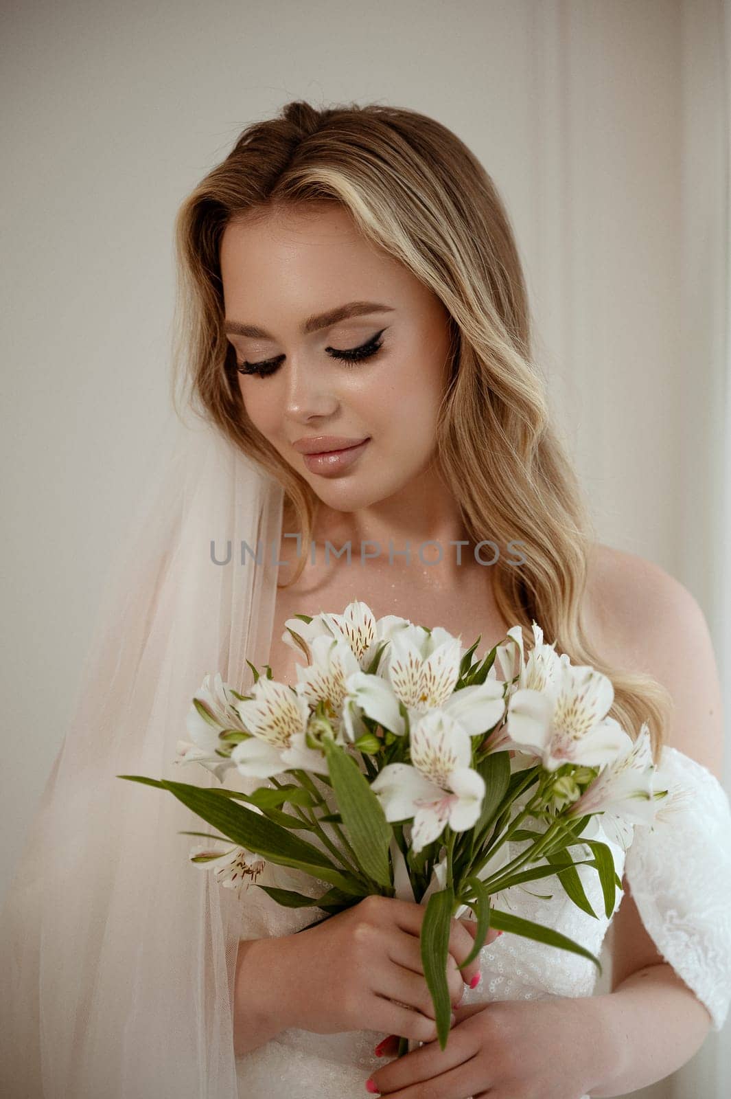 Portrait of a blonde bride with a lush veil, in a white dress with a bouquet of lilies. Close-up on a white background. Bride with makeup and wavy long hair, smiling