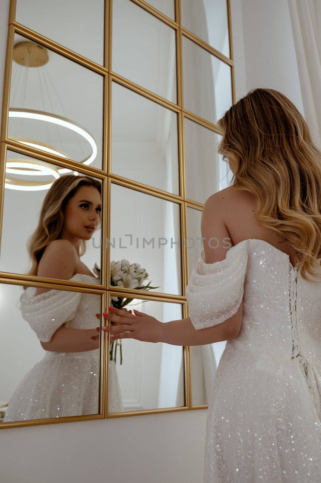 Bride with a bouquet of white flowers looks in the mirror. view from the back, blonde in a flowing wedding dress. with long hair, perfect hairstyle and makeup, smiling happily