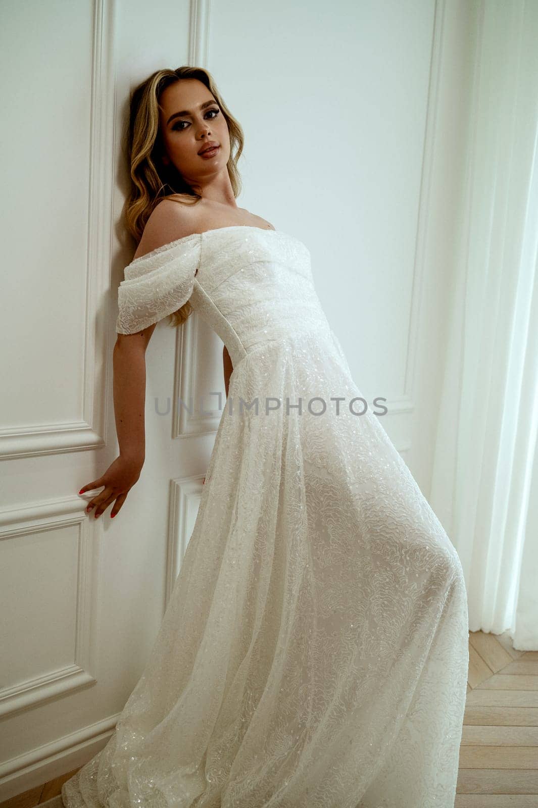 A stunning bride poses elegantly in a flowing wedding dress on a white background in a minimalist interior. She radiates grace and beauty. Blonde with long hair poses elegantly. High quality photo