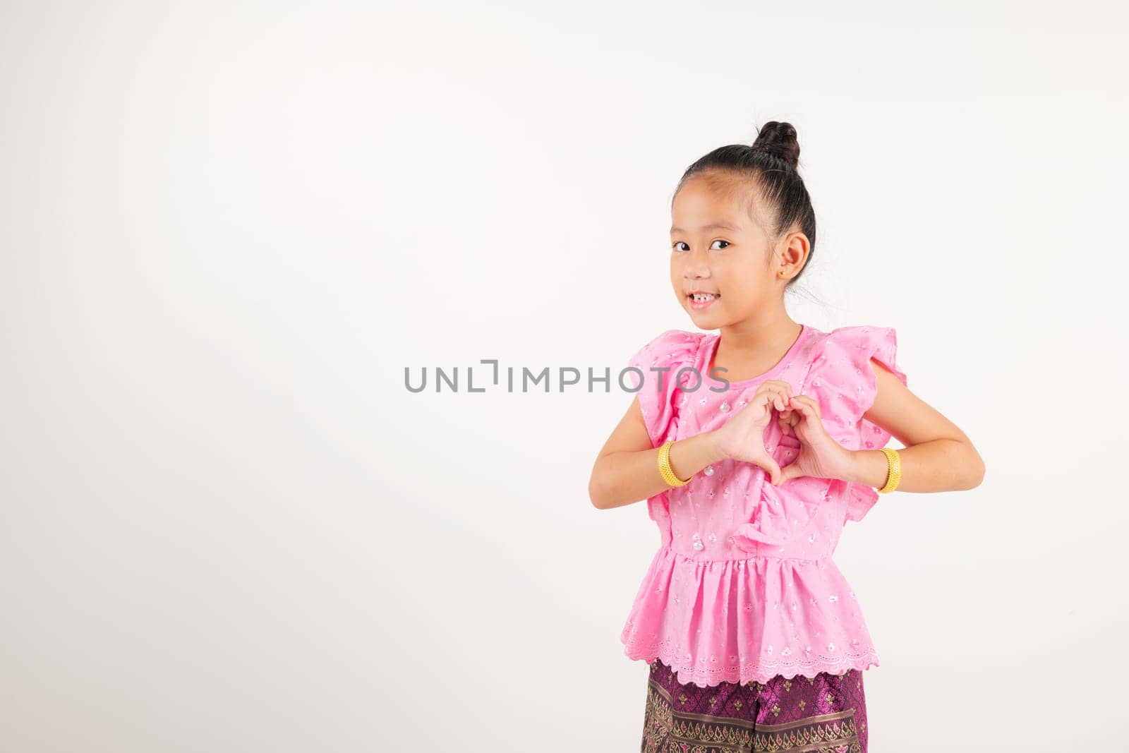 Portrait Thailand kid girl smiling traditional Thai dress costume shows creating heart shape with fingers and hands, studio isolated white background, kindergarten child sending love for Valentine day