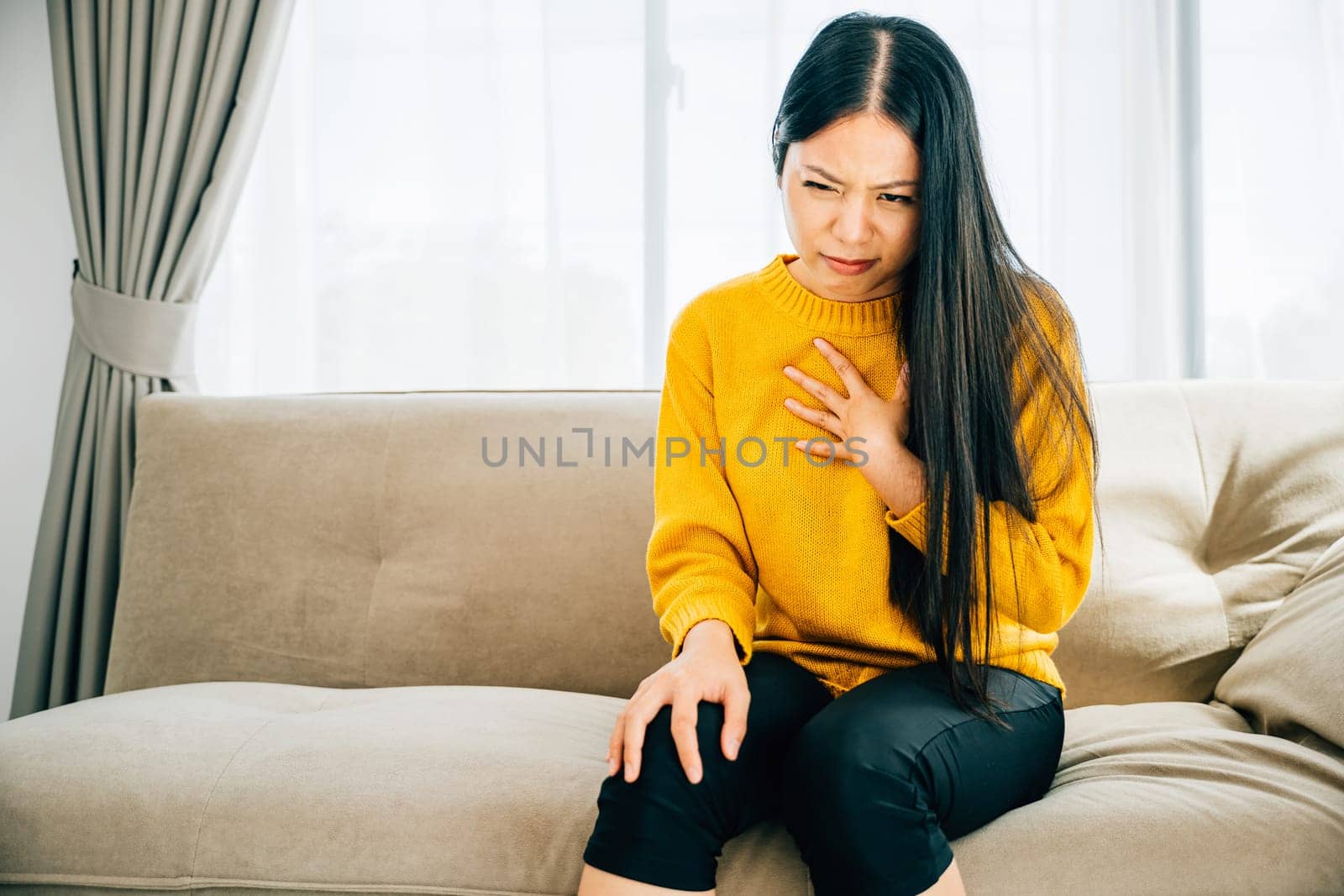 Highlighting a heart attack, Asian woman on a sofa clutches her left chest in pain needing immediate medical attention. Illustrating heartache discomfort and the urgency for emergency aid.