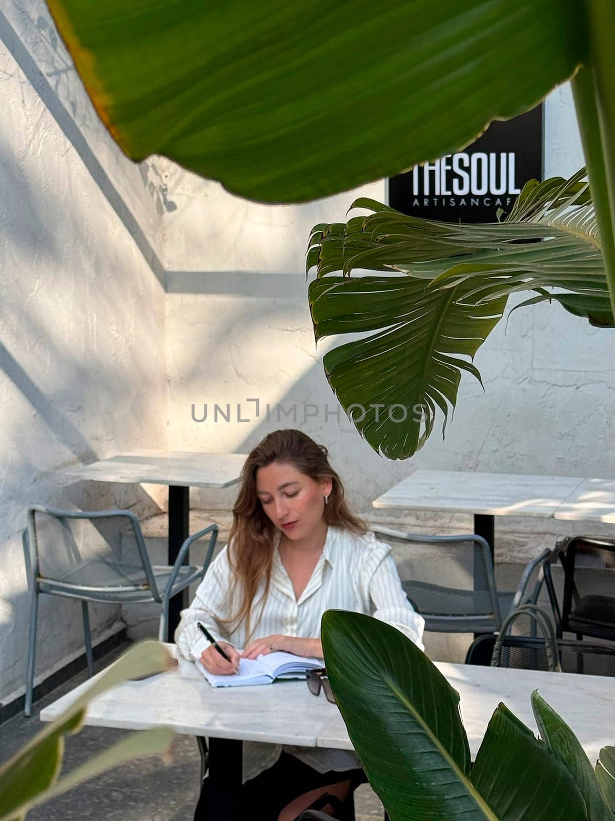 A young woman focuses on writing in her notebook at an outdoor cafe surrounded by leafy plants on a sunny day.