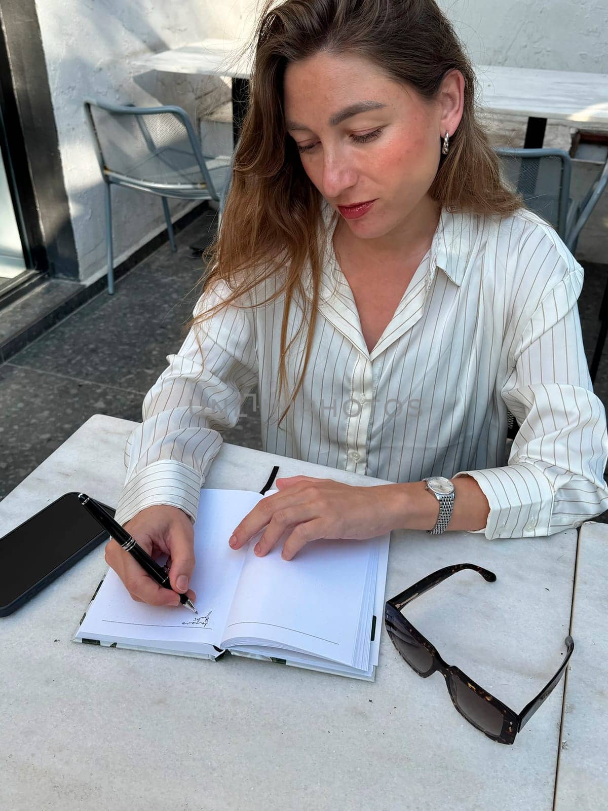 A woman in a striped shirt is focused on writing in a notebook at a stylish coffee shop table. Sunglasses rest nearby.