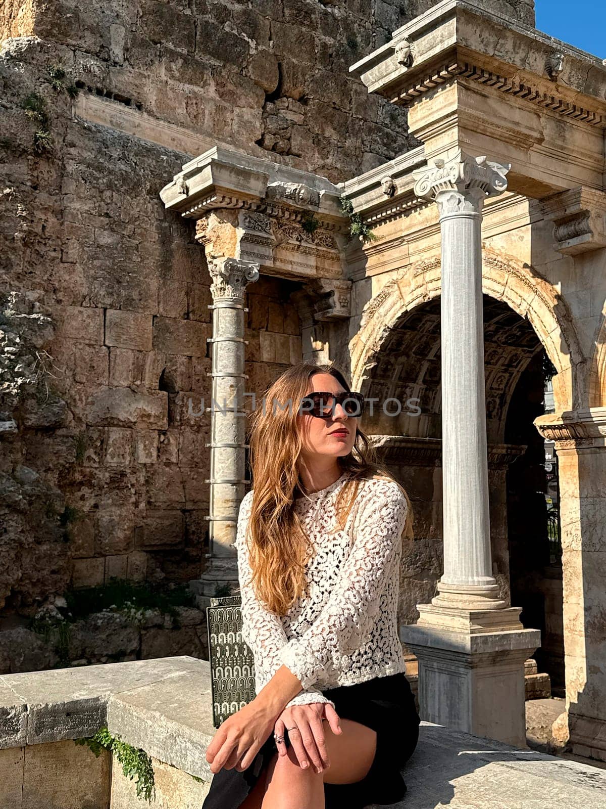 A woman sits elegantly on a stone ledge near ancient ruins, basking in the sunlight under a clear blue sky.