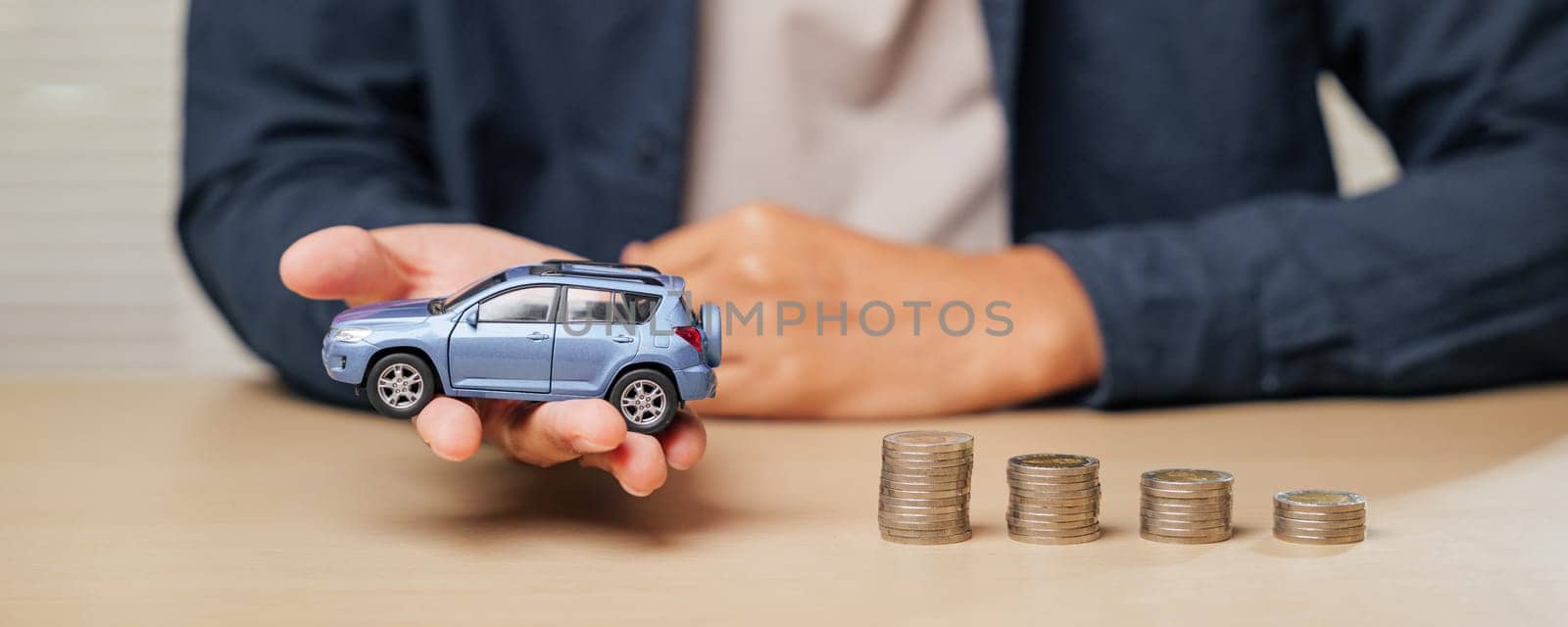 Hand holding a blue toy car model next to ascending coin stacks. Depicts auto credit, car loans, cash installment payments, and financial planning for managing car purchase costs.