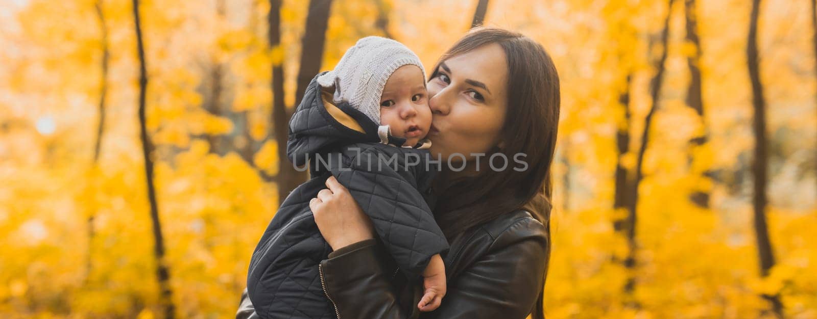 Autumn scene of mother kissing young baby girl outdoors with colorful trees in park