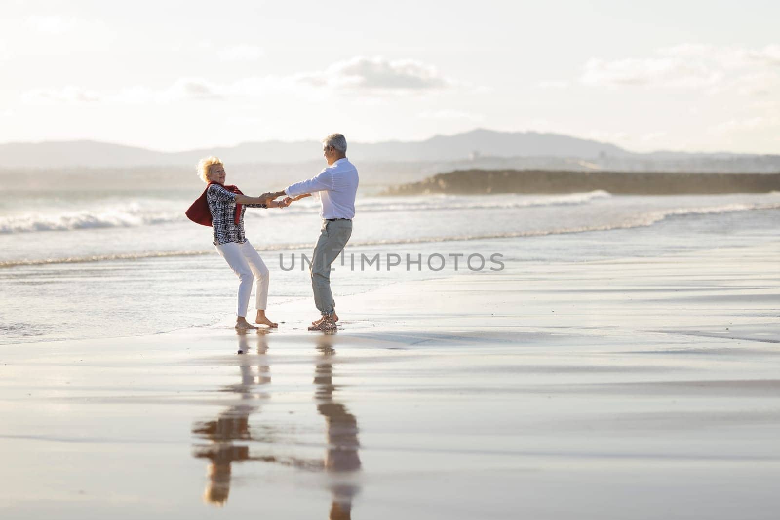 Elderly couple holding hands and spinning around on a wet sandy beach at sunset