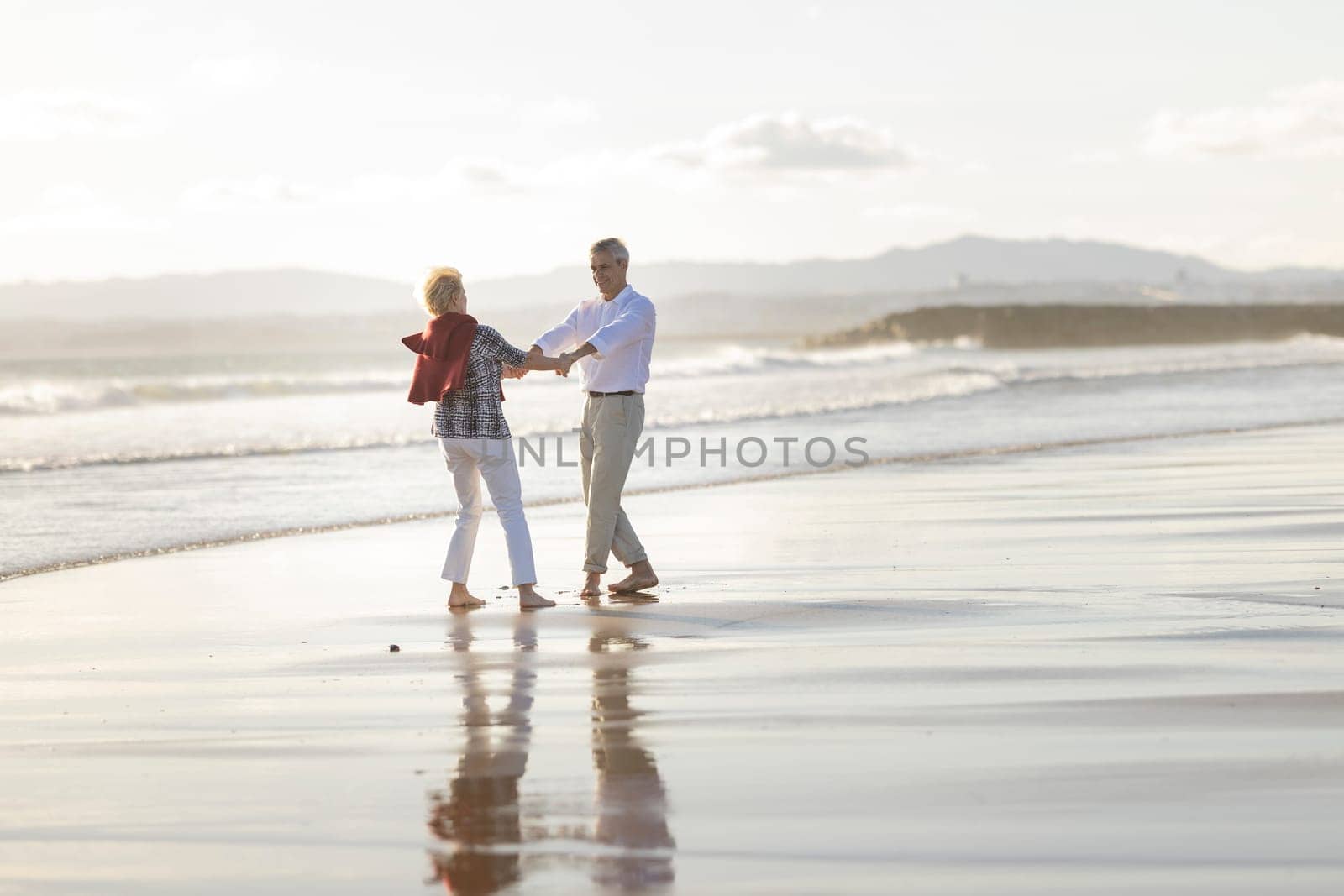 Carefree senior couple holding hands and dancing together on the beach at sunset, enjoying retirement