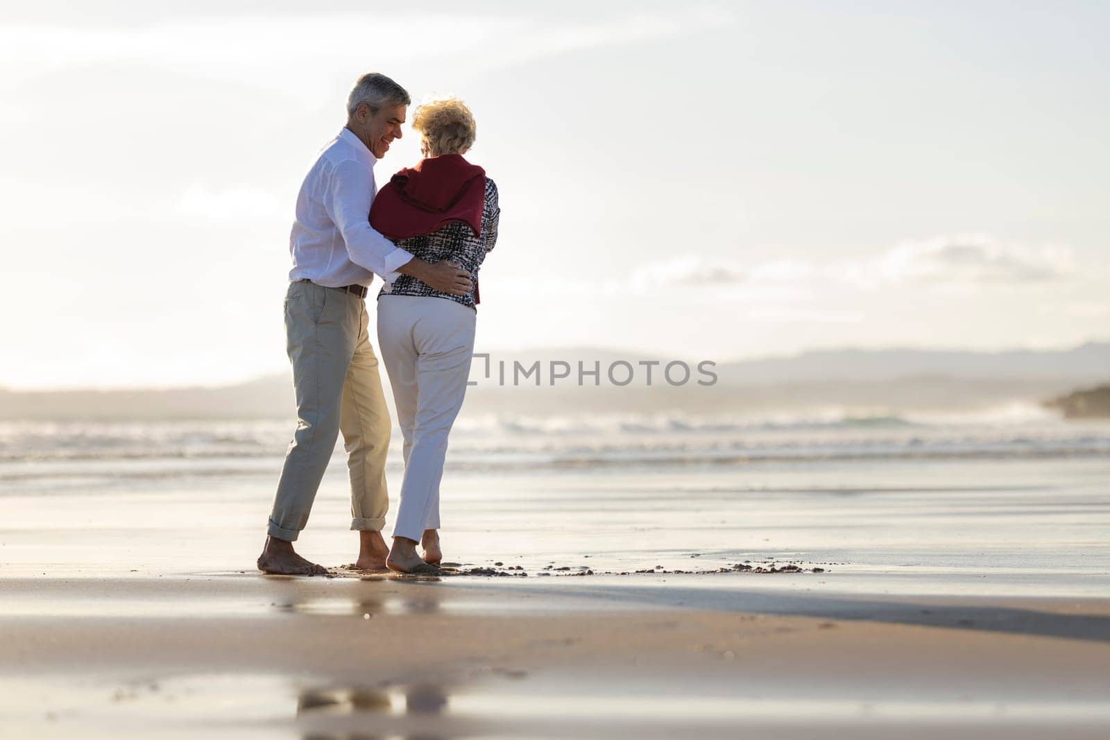 Happy retired couple embracing and walking on the beach at sunset, enjoying peaceful retirement