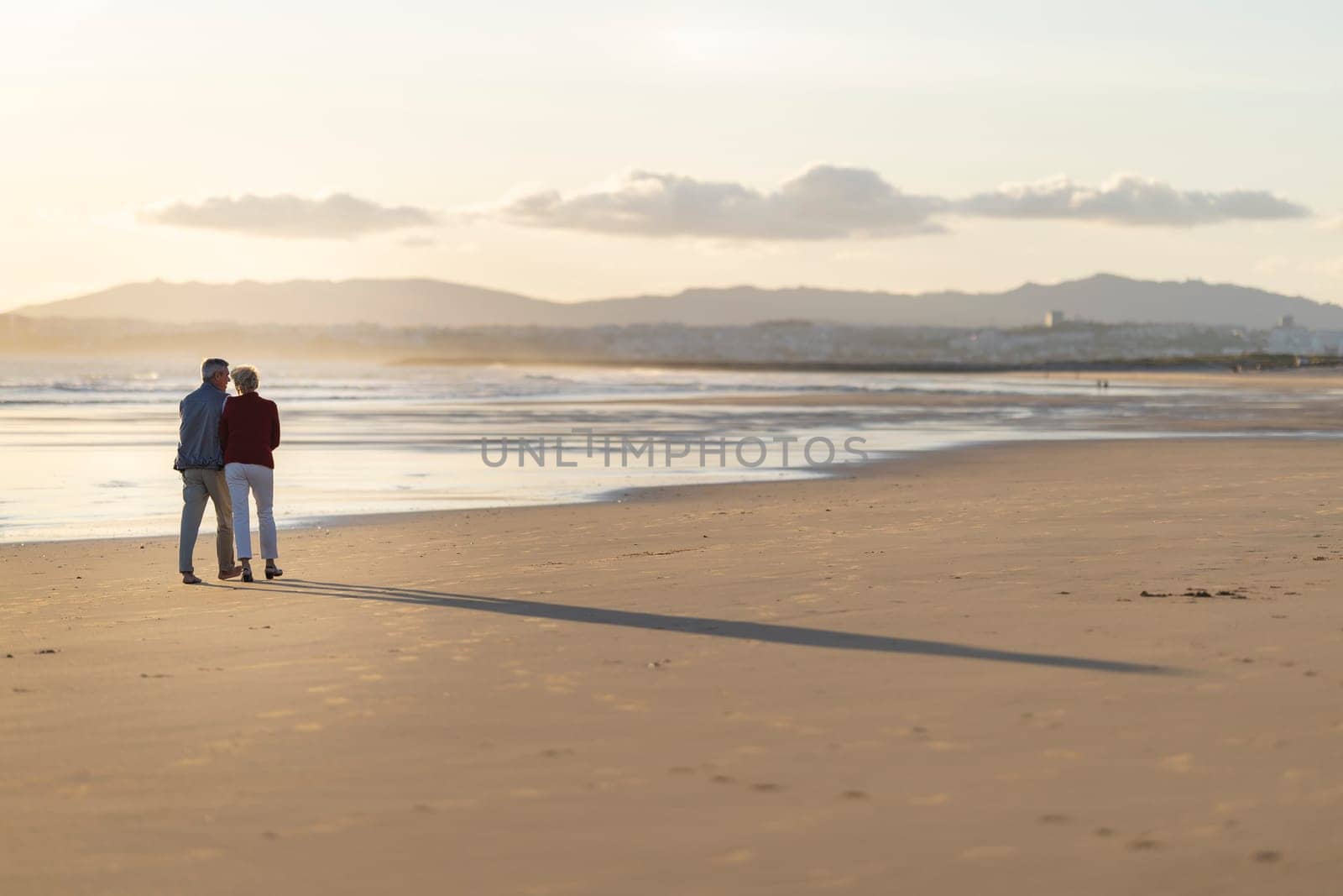 Elderly couple enjoying a romantic walk on the beach at sunset, creating a heartwarming scene of love and companionship