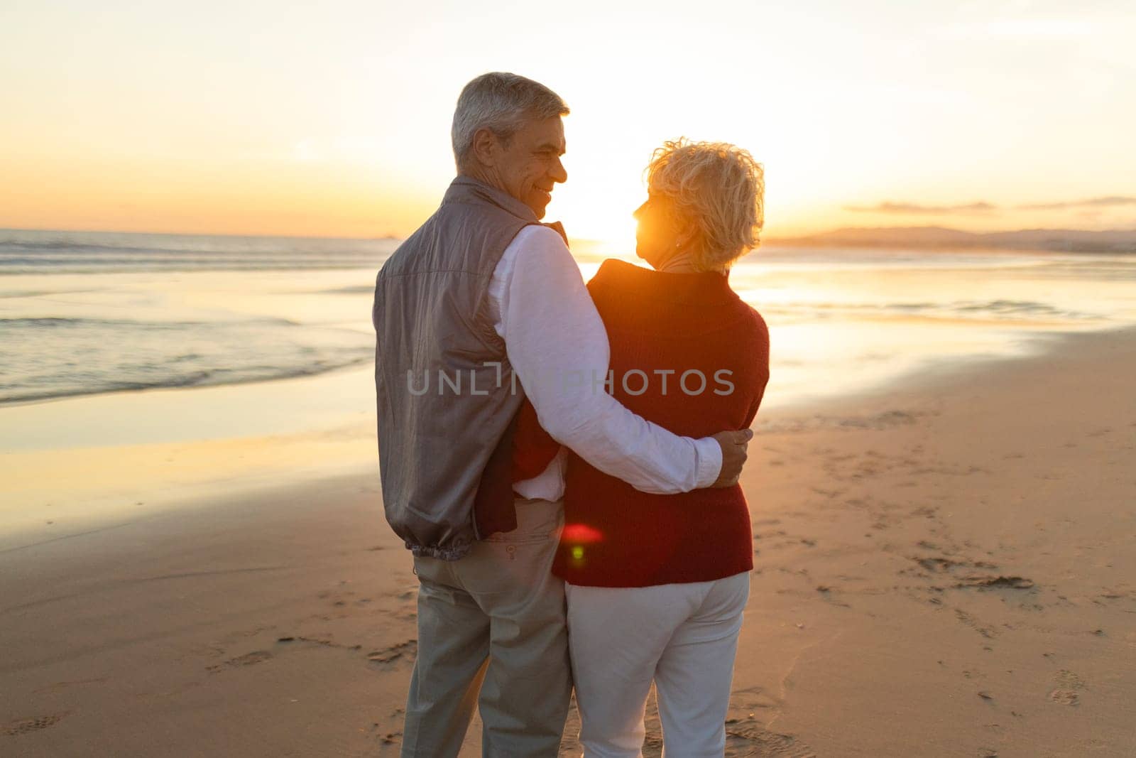 Elderly couple enjoying a romantic moment on the beach at sunset, embracing and looking at each other with love and affection