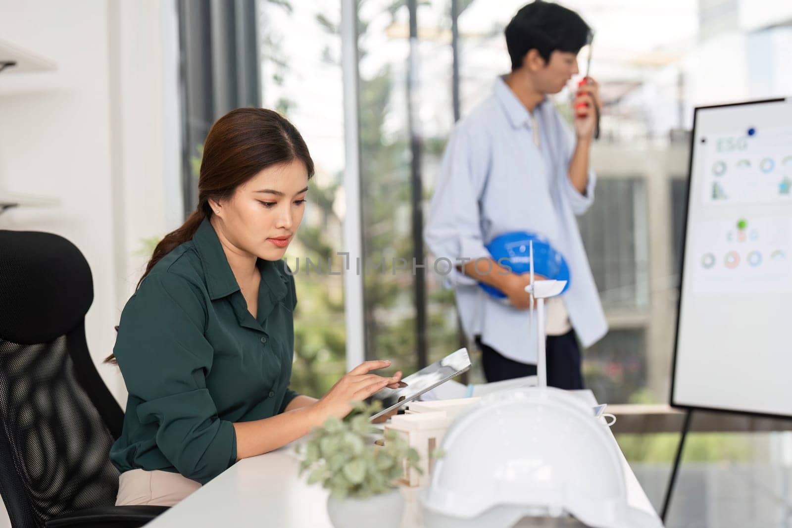 A female engineer attentively works on her tablet, analyzing project details while her colleague engages in the background, showcasing a collaborative workspace.