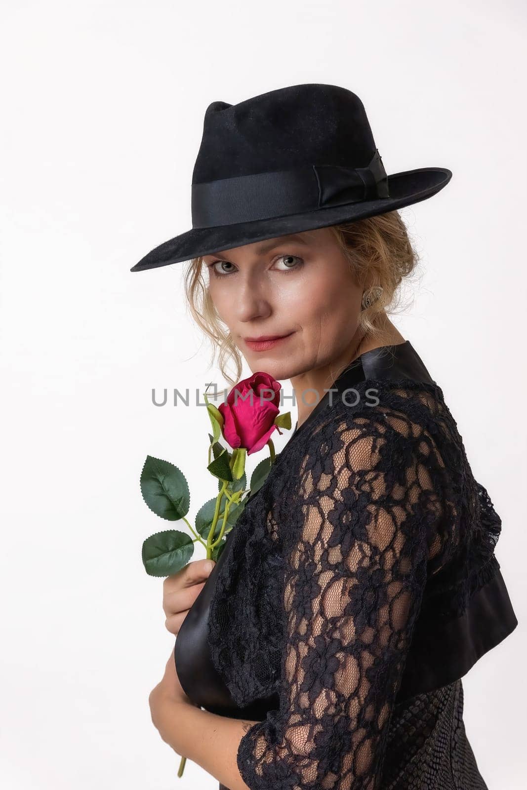 Middle aged woman in a black lace dress and hat holding a red rose in her hand is posing on a white background.