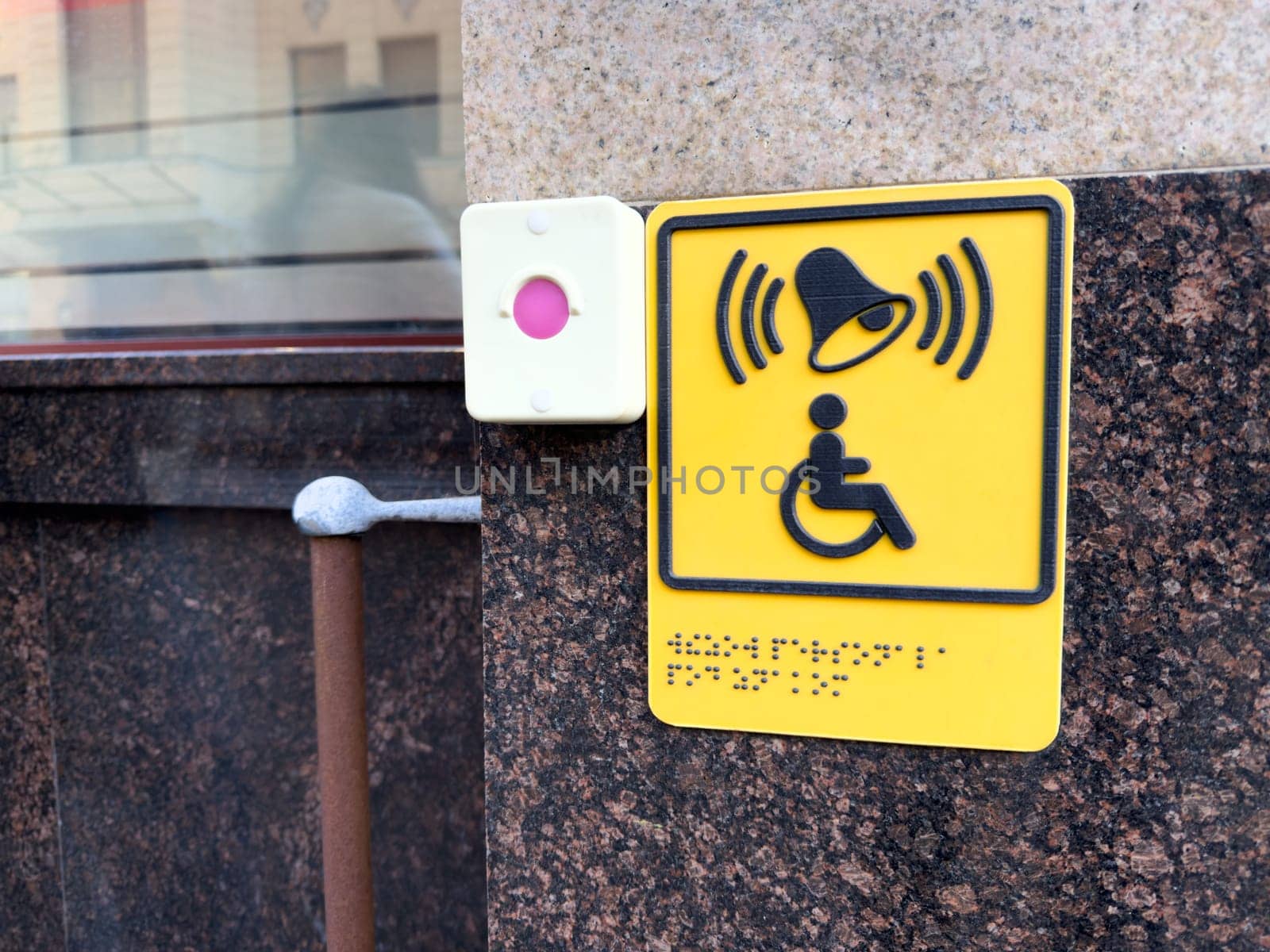 Bright yellow accessibility button for disabled assistance with Braille text and signal icon mounted on granite wall near entrance, promoting inclusive urban infrastructure
