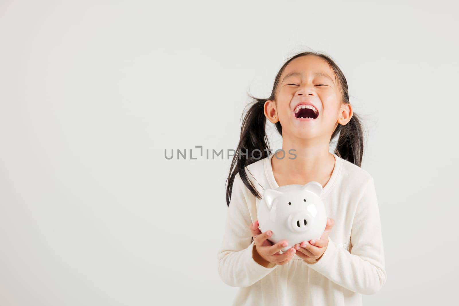 Asian little kid girl holding piggy bank and looking at camera at studio isolated on white background, Happy kindergarten child lifestyle smiling with is white piggybank, Thai personal money savings