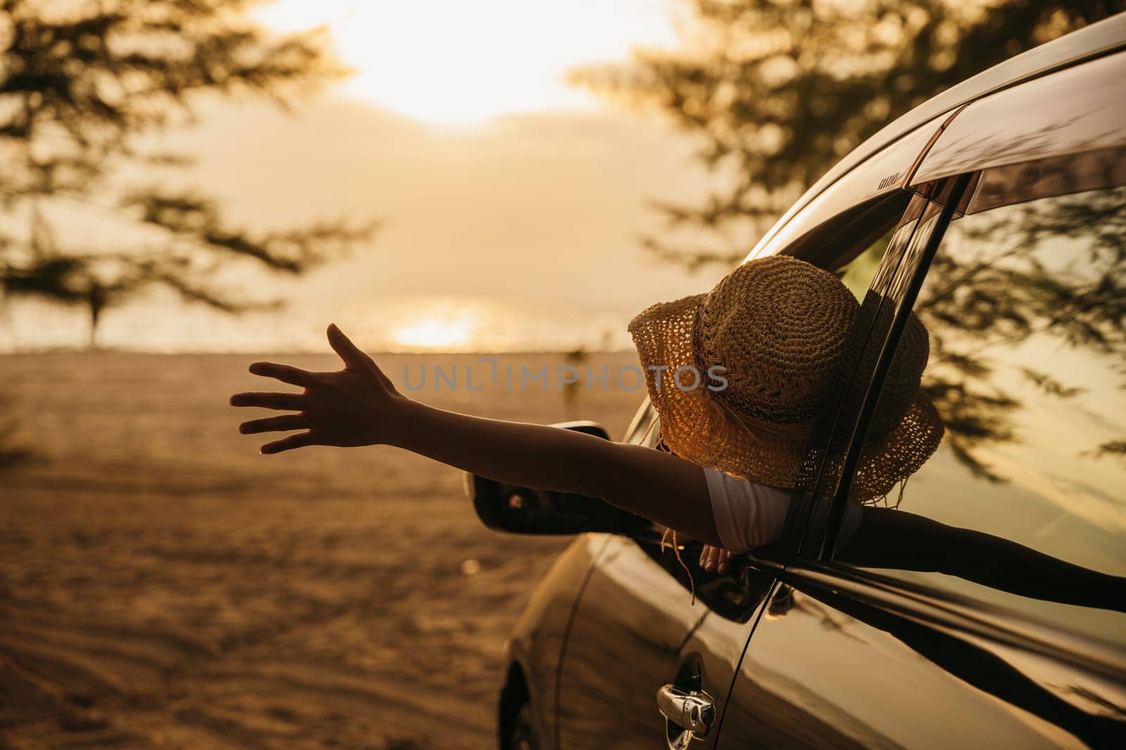 Woman wearing a stylish hat sits in front seat waving hand out car window at sunset feeling the wind. Happy weekend travel enjoy summer at beach. road trip and freedom concept
