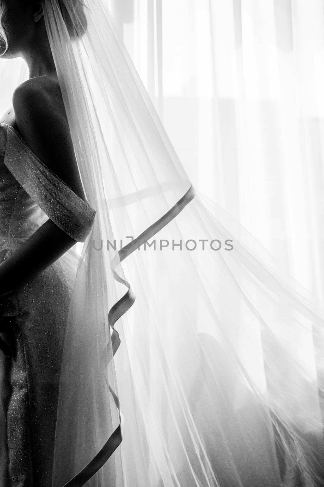 Silhouette of a blonde bride with a lush veil, in a long white dress near a window with a bouquet of lilies. Minimalistic interior, hotel or house.