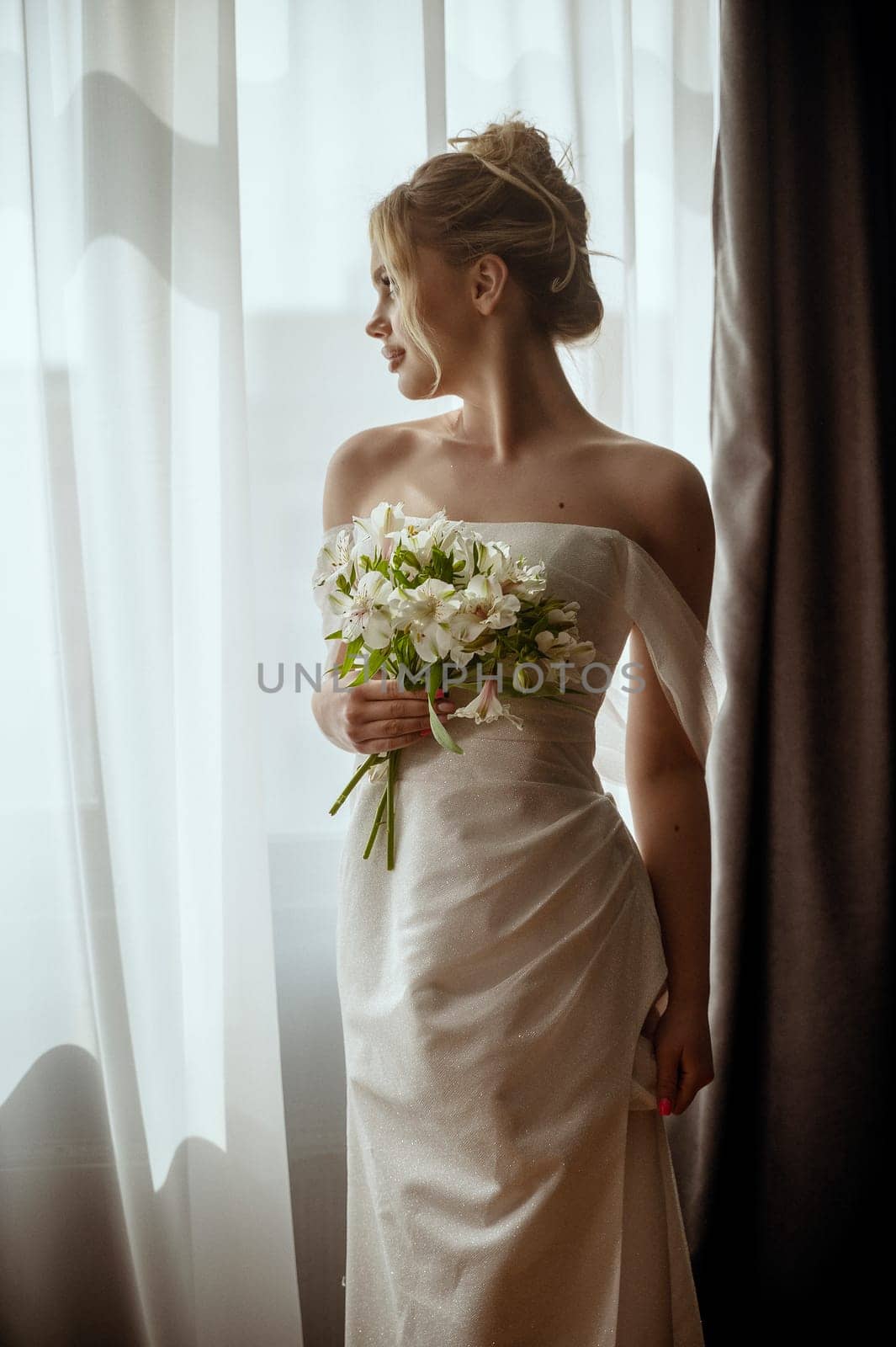 Beautiful blonde bride standing in a long white dress near the window with a bouquet of lilies in her hands. Minimalistic interior, hotel or house. bride in a long dress with a slit on the side