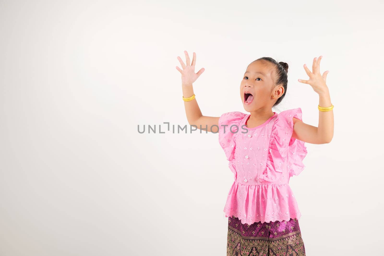 Portrait Thailand kid girl smiling traditional Thai dress costume raises fists in celebration of her success, studio isolated white background, kindergarten child saying yes with excitement winning