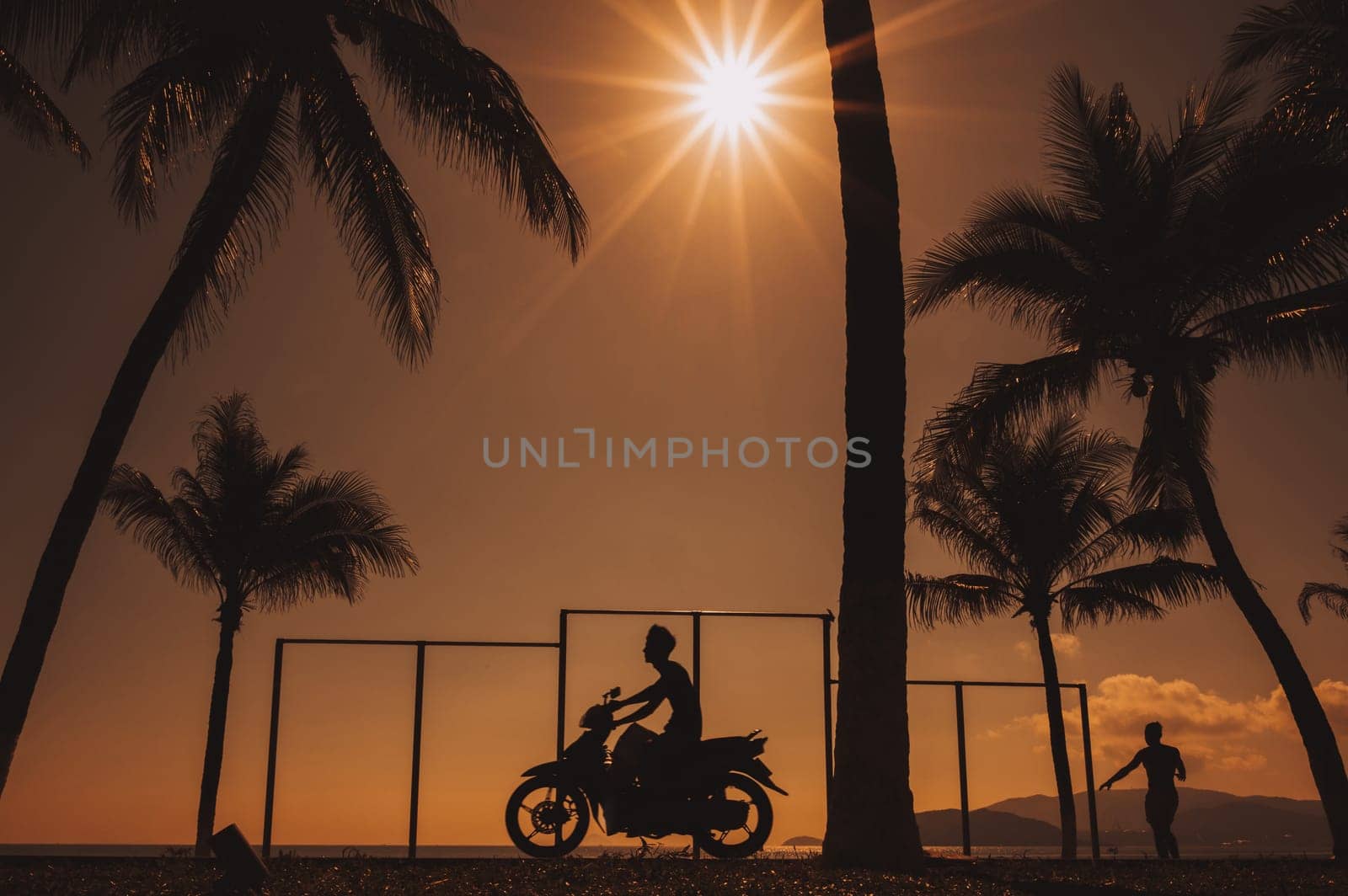 Silhouette of a Vietnamese man on a motorcycle on the embankment with palm trees by sea at sunrise on summer morning in Vietnam