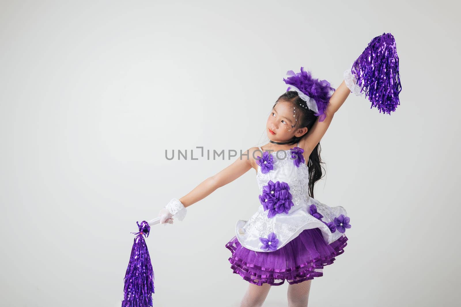 Young girl dressed in cheerleading gear, purple pom-poms in hand. Studio photo with white background, full of festive energy. cheerleader kid