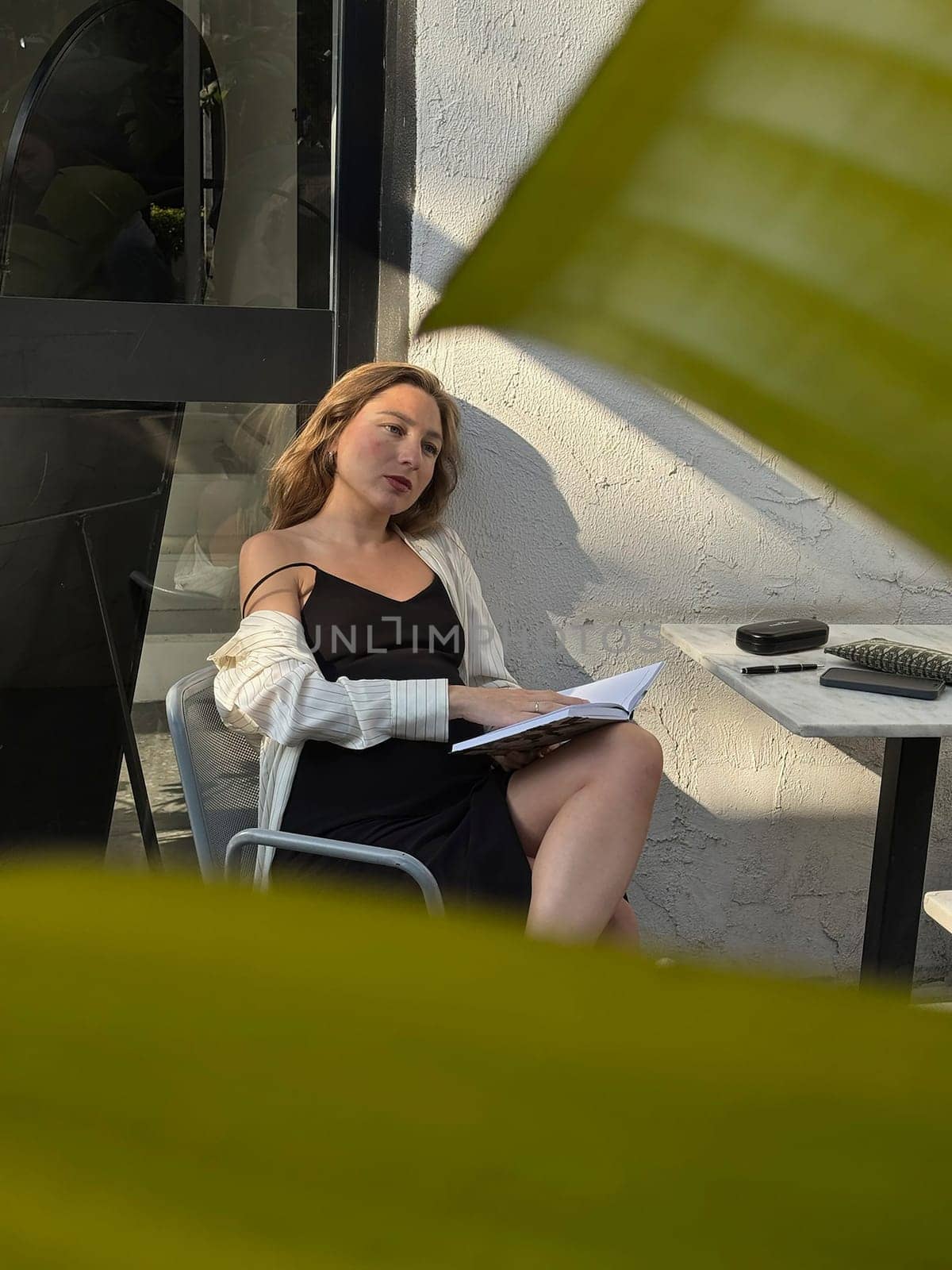 A woman sits relaxed at a table, reading a book while basking in the sunlight surrounded by green leaves.