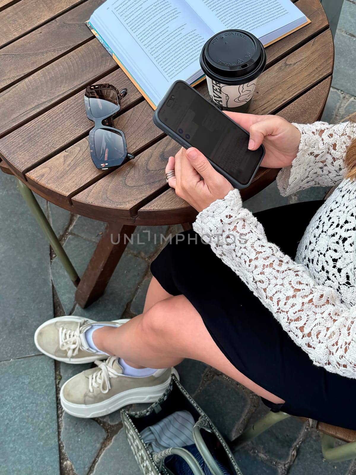 A person relaxes at a wooden table, sipping coffee and looking at their phone while reading a book in the afternoon.
