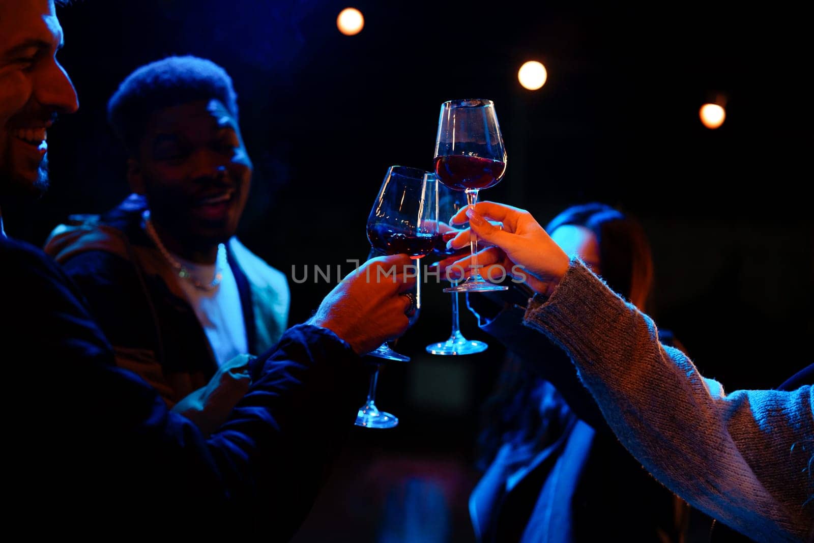 Group of friends enjoy a joyful moment together, clinking glasses filled with red wine under soft blue lighting at a vibrant nighttime gathering.