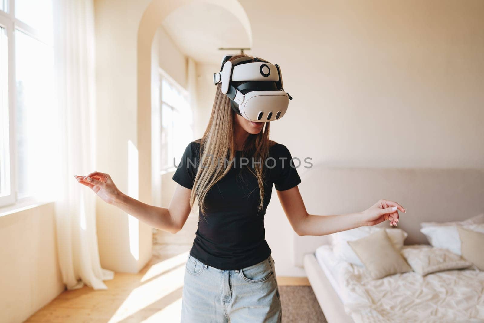 A young woman immersed in virtual reality gaming moves her hands gracefully while wearing a VR headset in a cozy, sunlit room. The background features light-colored furniture.