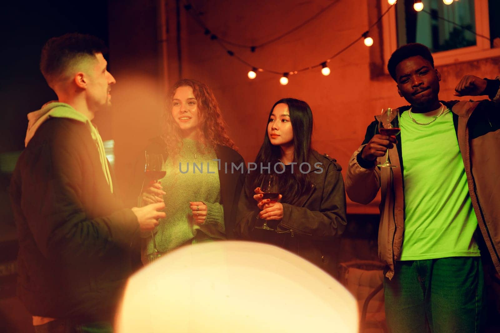 Group of four friends enjoy a lively evening outdoors sharing drinks and engaging in conversation. The vibrant atmosphere is enhanced by string lights illuminating their smiles.