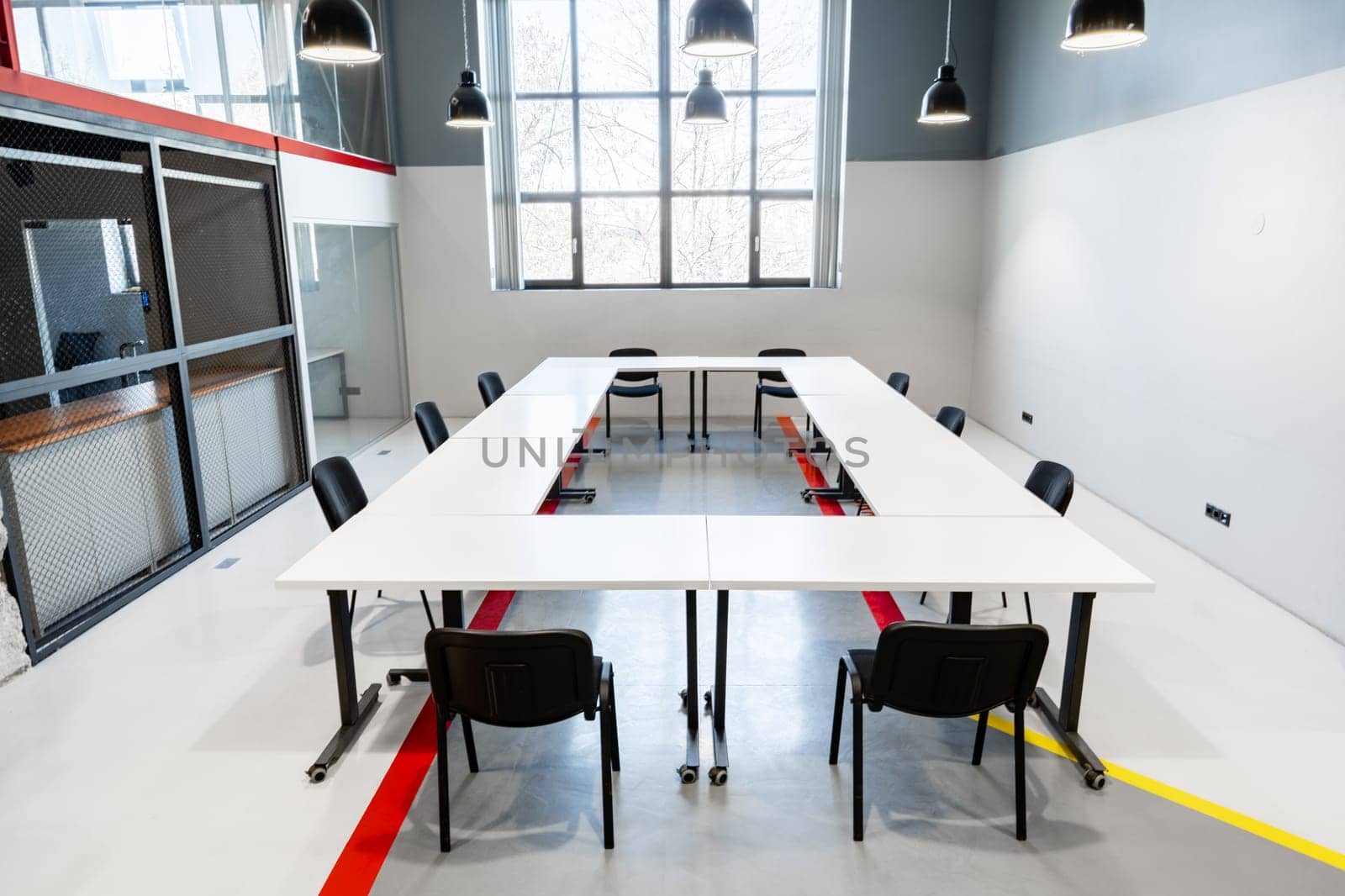 A spacious conference room features a long, rectangular table surrounded by black chairs. Industrial pendant lights hang from the ceiling, illuminating the minimalist design and large windows.