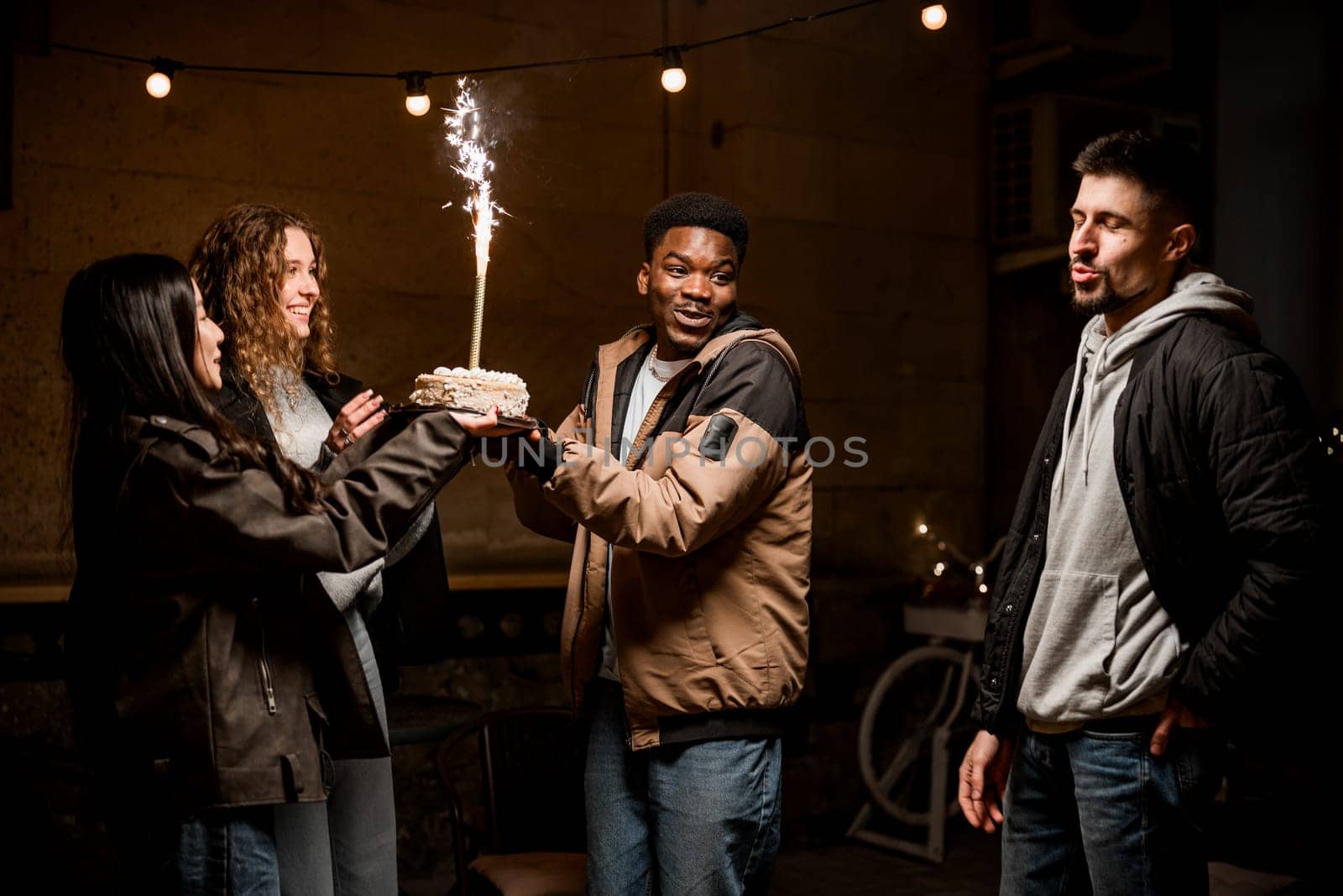 Friends celebrate together at an evening gathering, one person holding a cake adorned with a sparkling candle. Laughter and smiles fill the atmosphere, creating a lively moment.