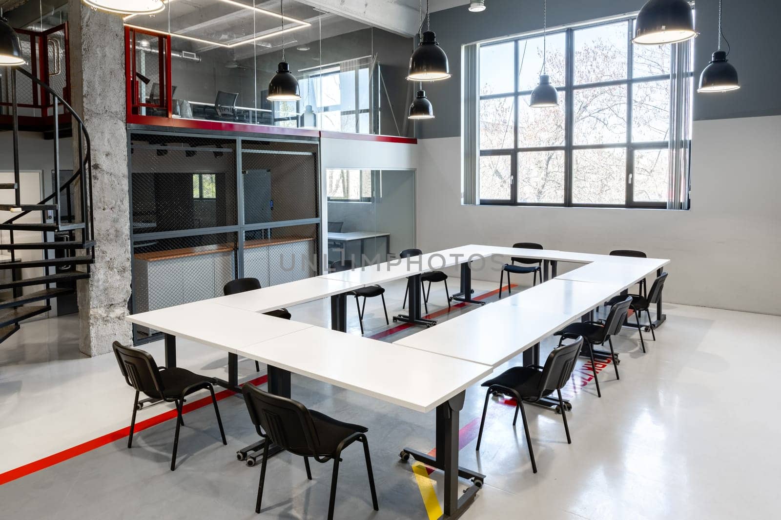 A spacious coworking area features a large white table surrounded by chairs. Large windows let in natural light, enhancing the modern design and atmosphere suitable for teamwork.