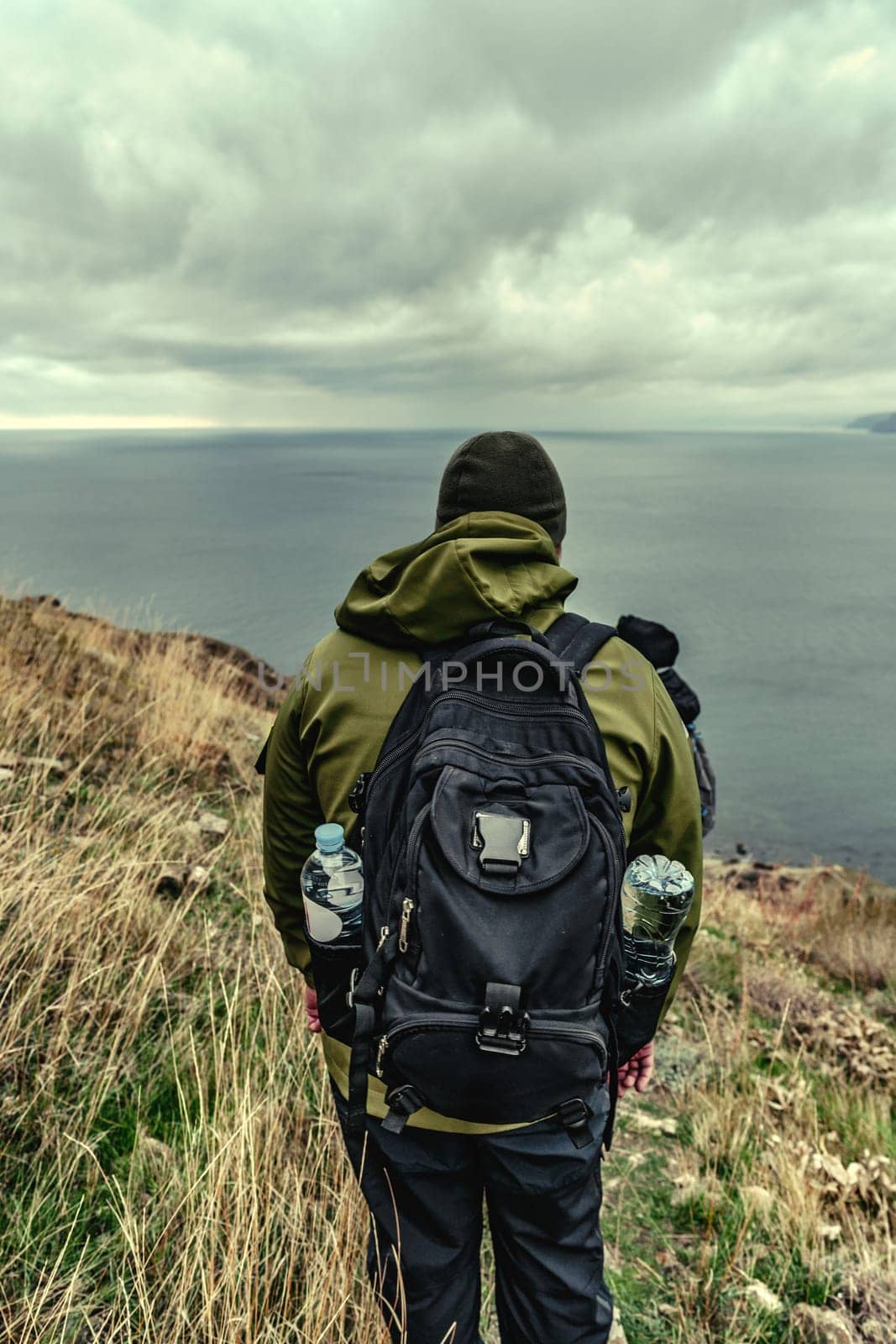 A lone hiker stands on a grassy cliff overlooking the ocean. Dressed in warm clothes, the person surveys the expansive seascape on a cloudy day.