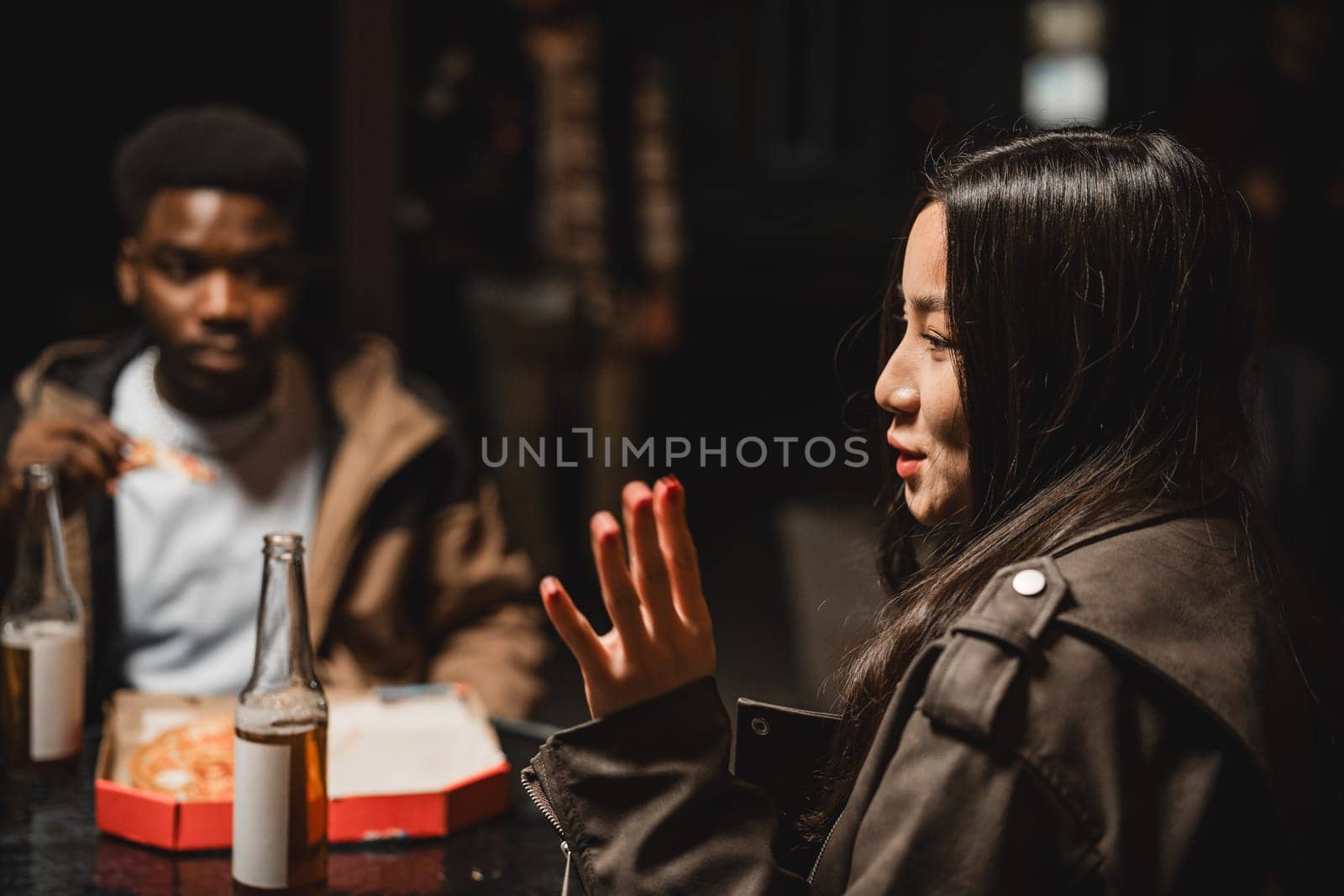 Two friends share a meal at a dimly-lit table, engaging in lively conversation while enjoying drinks and takeaway food in an urban environment.