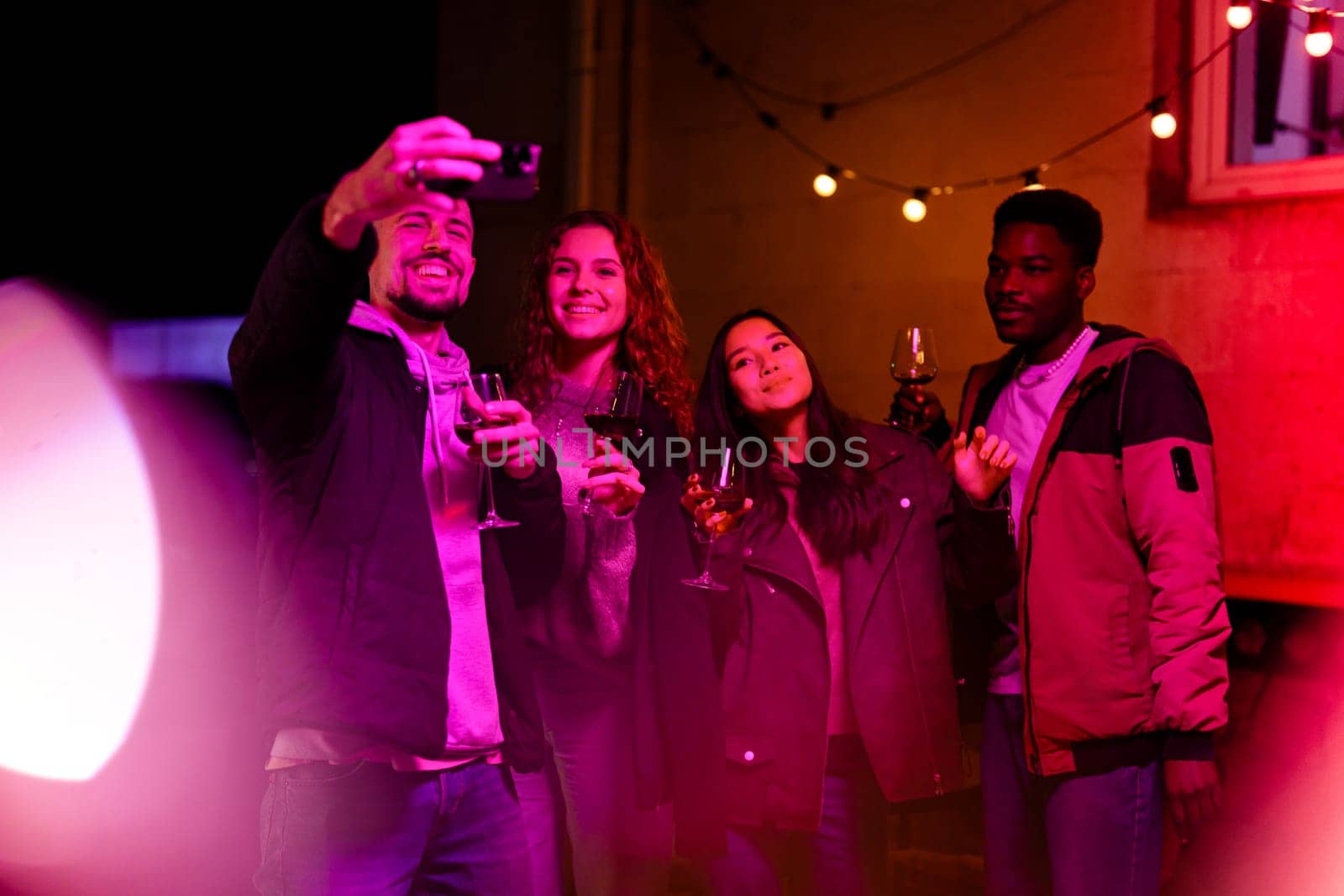 A group of four friends with drinks pose for a selfie in a cozy outdoor setting illuminated by string lights. Their joyful expressions capture the fun atmosphere of their gathering.