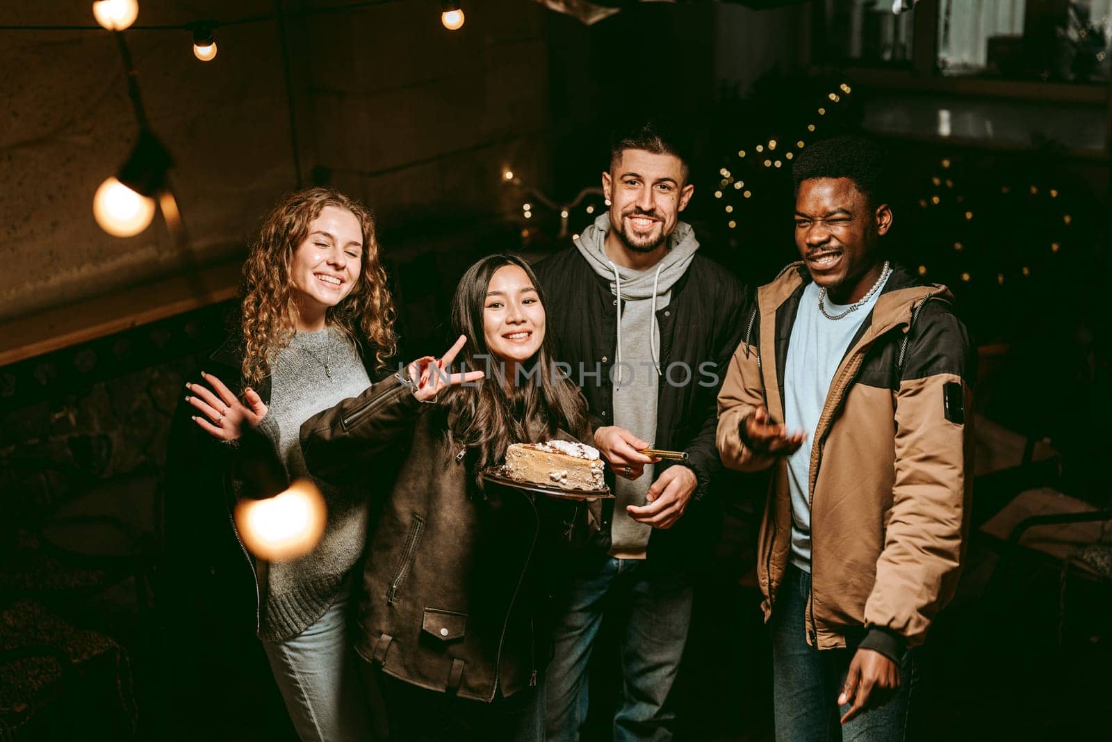 Four friends gather in a warmly lit venue, smiling and posing with a cake. The atmosphere is festive, filled with string lights and joyful expressions.