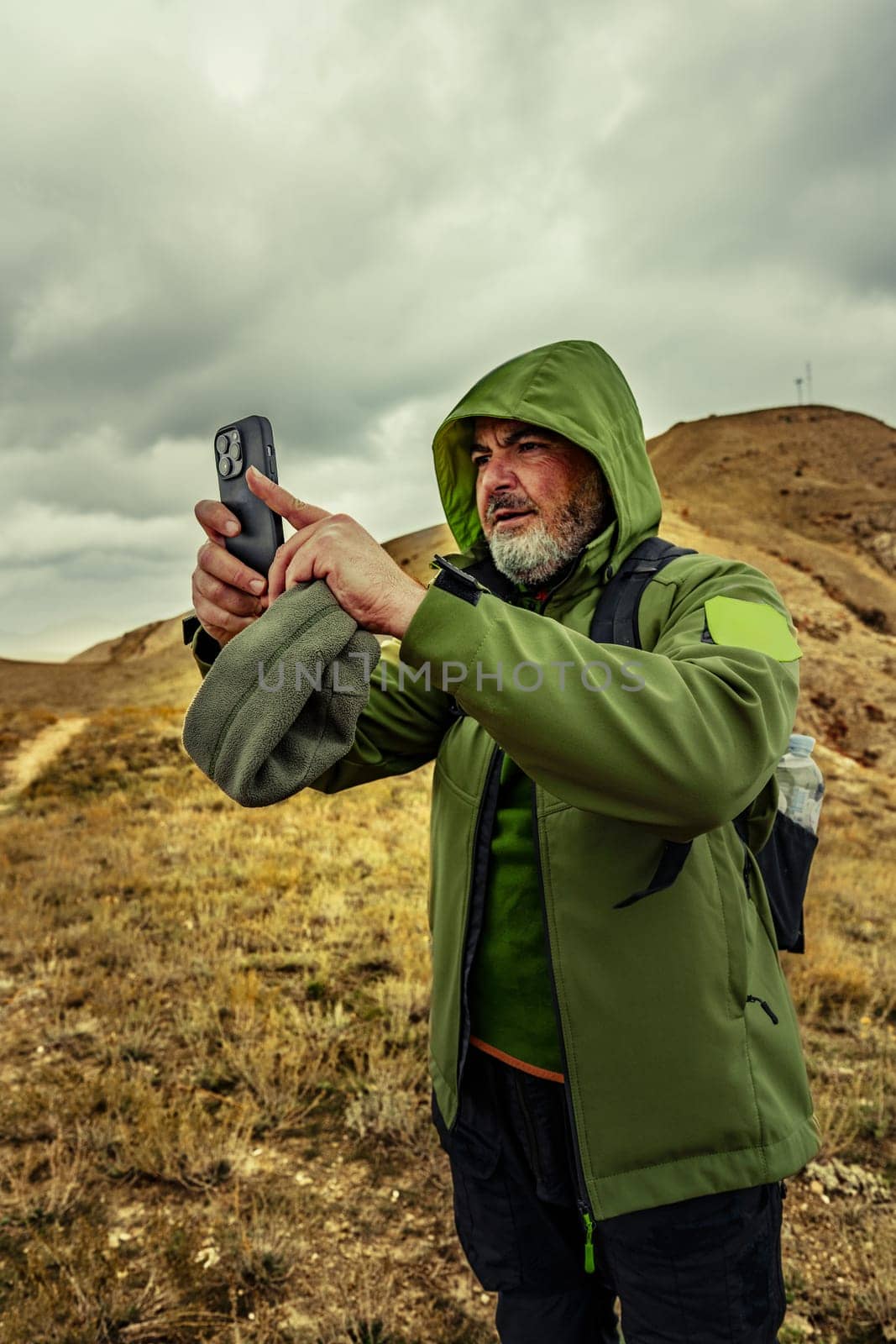 A man dressed in a green jacket takes a smartphone photo while hiking. The landscape features rolling hills and a dramatic clouded sky, showcasing a perfect outdoor adventure spot.