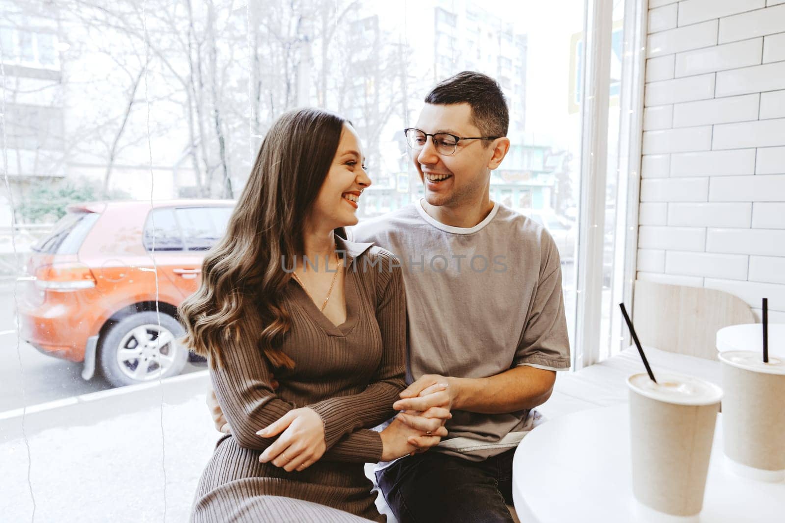 A couple sits closely together at a cafe, laughing and sharing a happy moment. They enjoy their drinks while sunlight filters through the windows, creating a warm atmosphere.