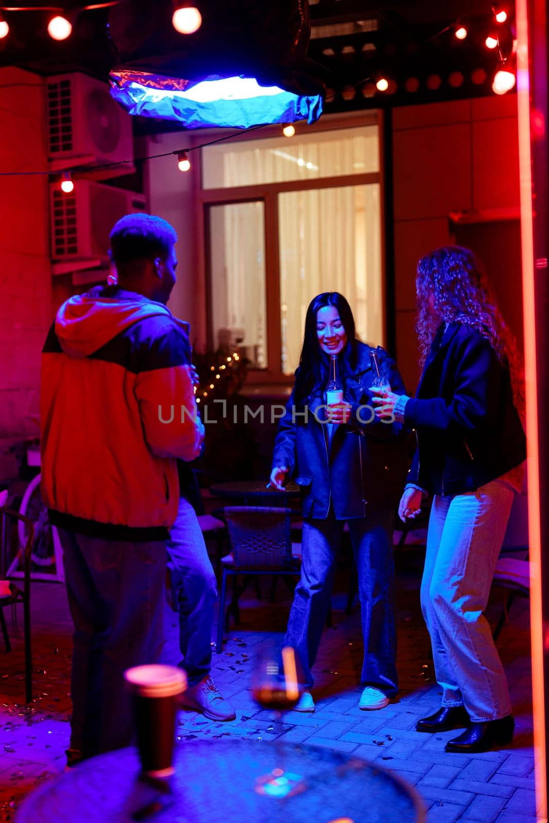 A group of three friends celebrates under vibrant lights, laughing and dancing together at an outdoor gathering. The atmosphere is lively, showcasing their joy and excitement.