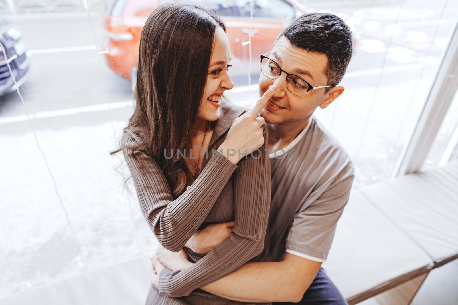 Inside a cozy cafe, a couple enjoys a light-hearted moment together. The woman playfully touches the man's nose while they smile at each other through a sunny window.