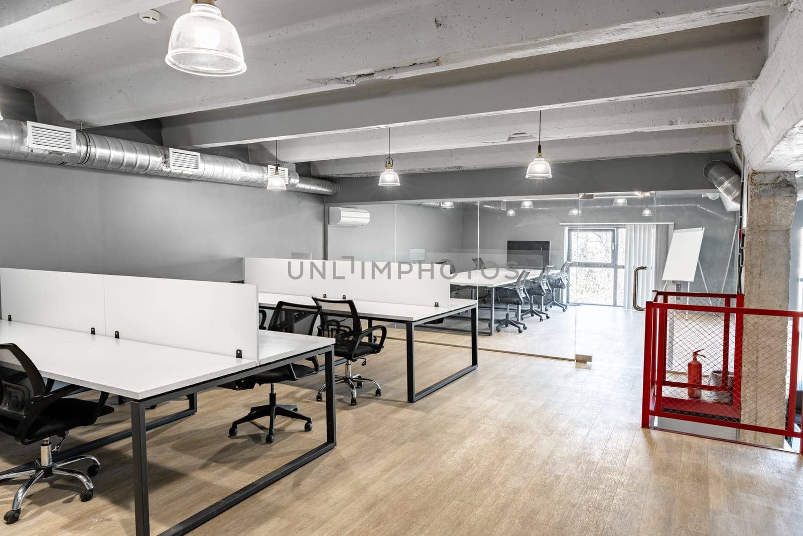 An open coworking area features several desks with ergonomic chairs in a spacious, well-lit setting. Large windows provide natural light, enhancing productivity.