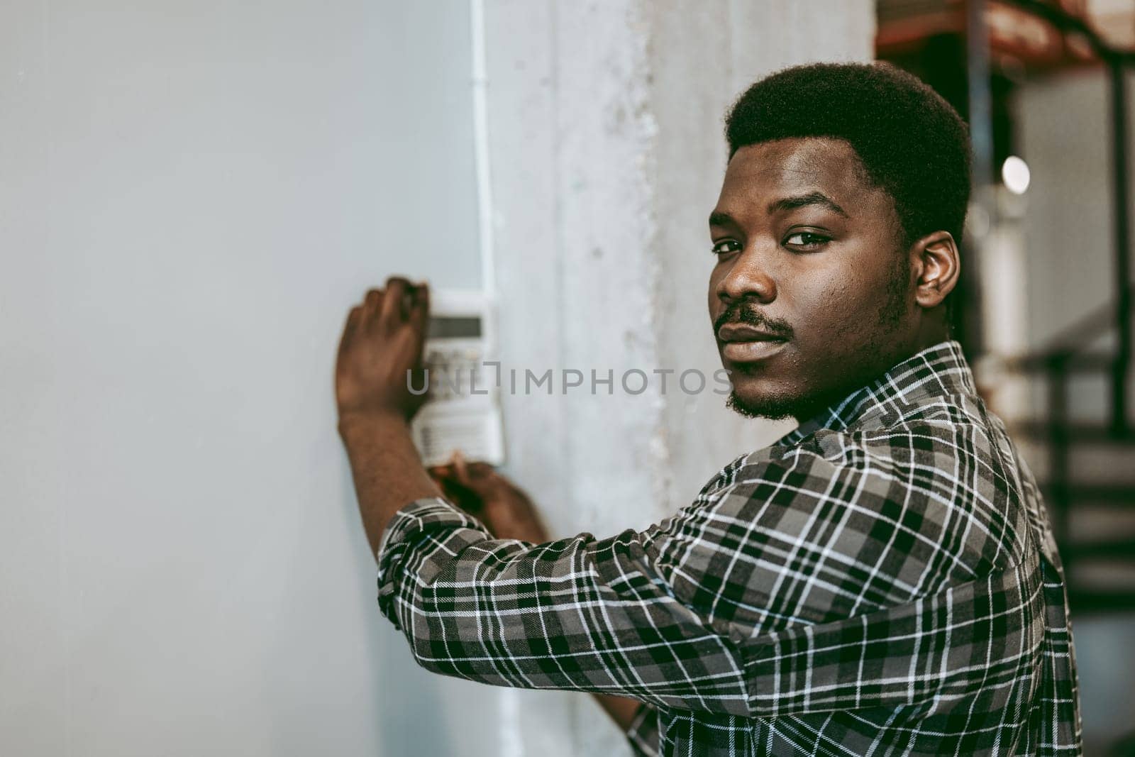 A man wearing a plaid shirt is focused on adjusting a device mounted on the wall. He is in a modern industrial setting, illuminated by natural light.